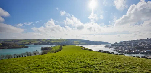 Snapes Point headland This is a landscape photograph of Snapes Point headland in Devon, England, United Kingdom, taken during the afternoon in early spring. The image prominently features the green grassy fields of Snapes Point extending towards the estuary, with the water of the Kingsbridge Estuary clearly visible. In the background on the right, the town of Salcombe can be seen nestled along the estuary’s edge, with its cluster of buildings adding detail to the coastal scene. The sky is partly cloudy with the sun high, casting bright light across the landscape, emphasizing the fresh spring vegetation on the headland. The photograph captures the characteristic countryside of Devon with rolling hills and waterways under clear, seasonal lighting.